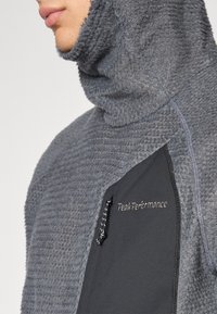 Grå fleece hettegenser med strukturfinish, med en svart sidepanel og en brystlomme med snøring. Logo vist på brystet.