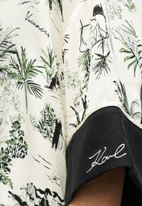 Κοντομάνικο πουκάμισο με λευκή βάση, μαύρο τροπικό μοτίβο και πράσινες λεπτομέρειες. Διαθέτει μαύρο περίγραμμα και κεντημένη υπογραφή "Karl".