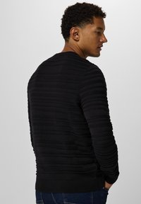 Schwarzer gestrickter Pullover mit geripptem Muster und horizontalen Streifen, runder Ausschnitt und langen Ärmeln, getragen mit blauen Jeans.