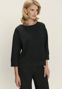 Mujer con cabello rubio vistiendo una blusa larga de manga negra texturizada y pantalones a juego, de pie frente a un fondo neutro, mirando hacia la derecha.