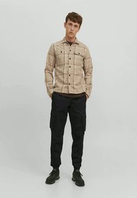 Camisa xadrez bege com fecho de botão, dois bolsos no peito; combinada com calças cargo pretas e sapatilhas pretas. Design simples e funcional.