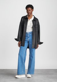 Chemise oversize en cuir noir, polo rayé blanc en dessous, jeans larges bleus et baskets blanches sur un fond neutre.