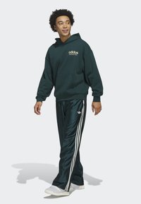 Tmavě zelená mikina s klokaní kapsou, na níž je zlatý nápis "adidas". V kombinaci se lesklými tmavě zelenými tepláky se třemi bílými pruhy a bílými teniskami.