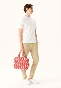 Rood patroon tas met witte accenten, rechthoekige vorm, dubbele handvatten en een rits. Model draagt een wit t-shirt en beige broek.