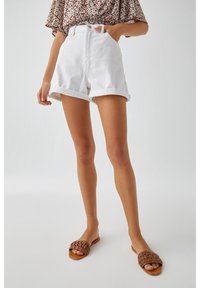 Femme portant un short blanc à ourlet roulé, un haut fleuri et des sandales tissées marron, debout devant un fond clair uni.