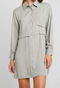 Ljusgrå, långärmad skjortklänning som är gjord av ett slätt tyg. Har en knappstängning fram, en bröstficka och en avslappnad passform med en lagerdesign.