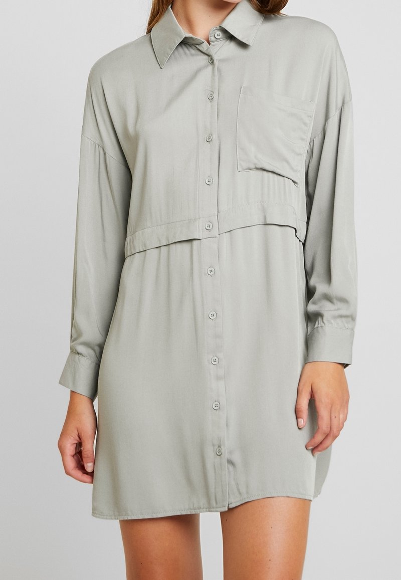 Ljusgrå, långärmad skjortklänning som är gjord av ett slätt tyg. Har en knappstängning fram, en bröstficka och en avslappnad passform med en lagerdesign.