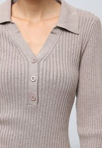 Haut en tricot côtelé beige avec un col en V et un col, comportant trois boutons assortis à l'avant. Texture douce, design ajusté et manches longues.