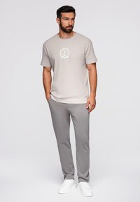 Man met een baard, gekleed in een beige T-shirt met een ankerlogo, grijze broek en witte sneakers, staand met zijn handen in zijn zakken tegen een witte achtergrond.