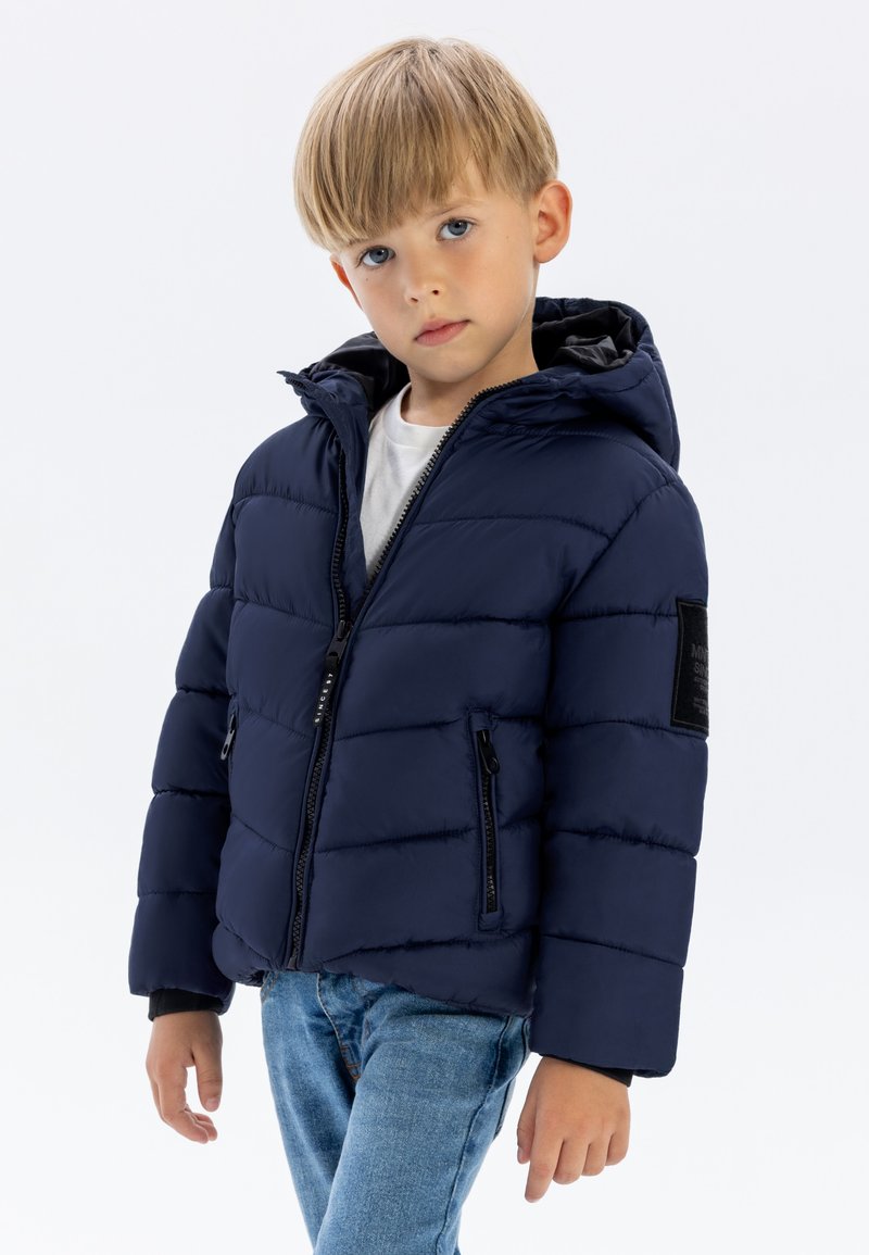 Navy padded jacket with a hood, front zipper, side pockets, and black fabric patch on the sleeve. Worn over a white shirt and jeans.