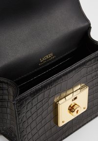 Svart läderhandväska med krokodilsmönster, försedd med ett guldmetallspänne och logotyp. Innerväggarna har en svart strukturerad foder.