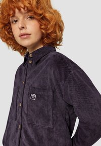 Donkerpaarse corduroy shirt met een knoopsluiting aan de voorkant, twee borstzakken en een klein geborduurd logo. Ribbelstructuur en puntige kraag.