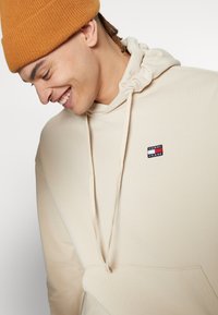 Beige Hoodie mit verstellbarem Kordelzug, ausgestattet mit einem kleinen blauen, roten und weißen Tommy Jeans Logo auf der Brust. Weiche, glatte Textur.