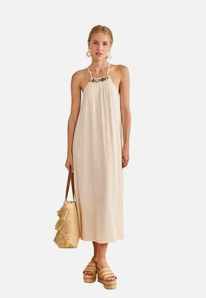 Mujer con un vestido midi sin mangas beige con cuentas de colores en el escote, sosteniendo un bolso tote de paja y calzando sandalias de plataforma tejidas.