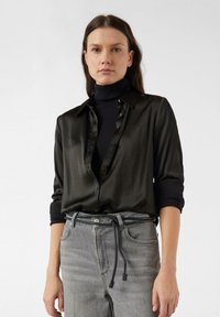 Czarna jedwabna koszula z kołnierzykiem na guziki, z podwiniętymi rękawami i gładką fakturą, noszona na czarnym golfie. Szare dżinsy ze sznurkiem w talii.