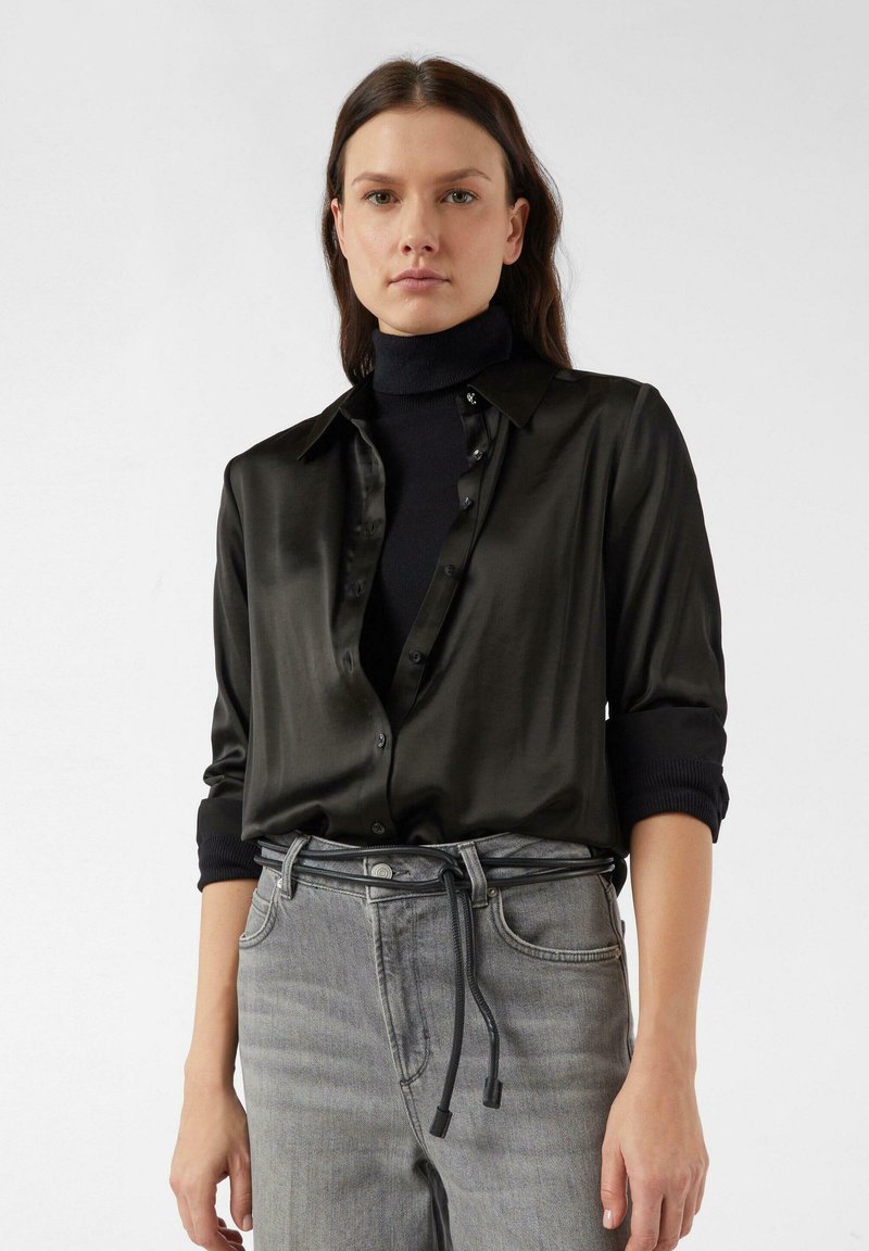 Czarna jedwabna koszula z kołnierzykiem na guziki, z podwiniętymi rękawami i gładką fakturą, noszona na czarnym golfie. Szare dżinsy ze sznurkiem w talii.