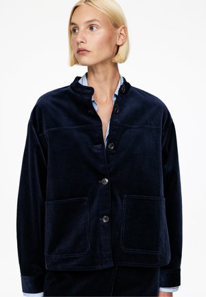 Frau mit kurzem blonden Haar, die eine dunkelblaue Cordjacke mit großen Vordertaschen und Knöpfen über einem hellblauen Hemd trägt.