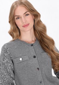 Cárdigan gris con mangas adornadas con lentejuelas, que presenta un cuello redondo, botones delanteros y dos bolsillos en el pecho. Textura suave y diseño ceñido.