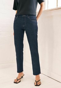 Jeans de mezclilla en azul oscuro con un ajuste slim, que cuentan con bolsillos frontales y detalles de costura sutiles. Combinados con sandalias negras.