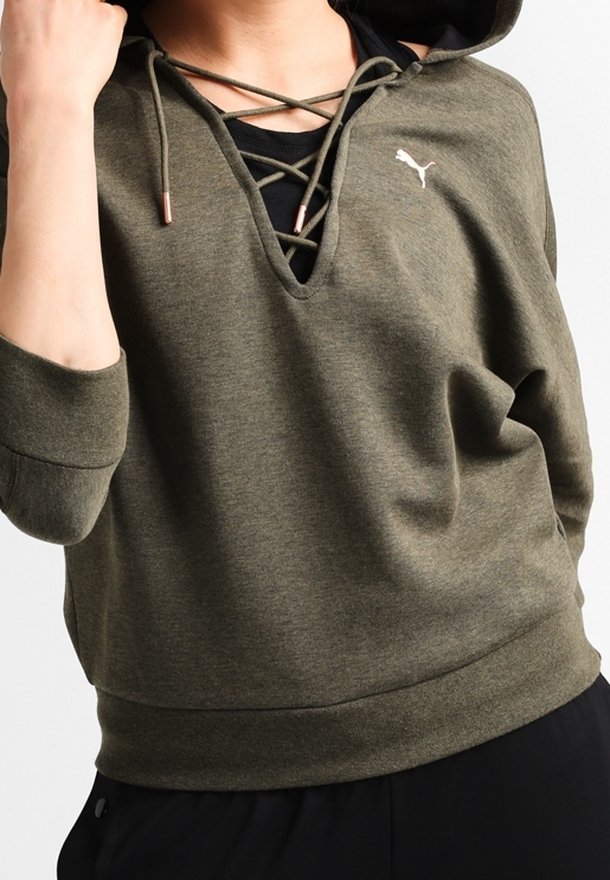 Personne portant un sweat à capuche Puma vert olive avec un col lacé et un t-shirt noir en dessous, vue recadrée montrant le torse et le bras.