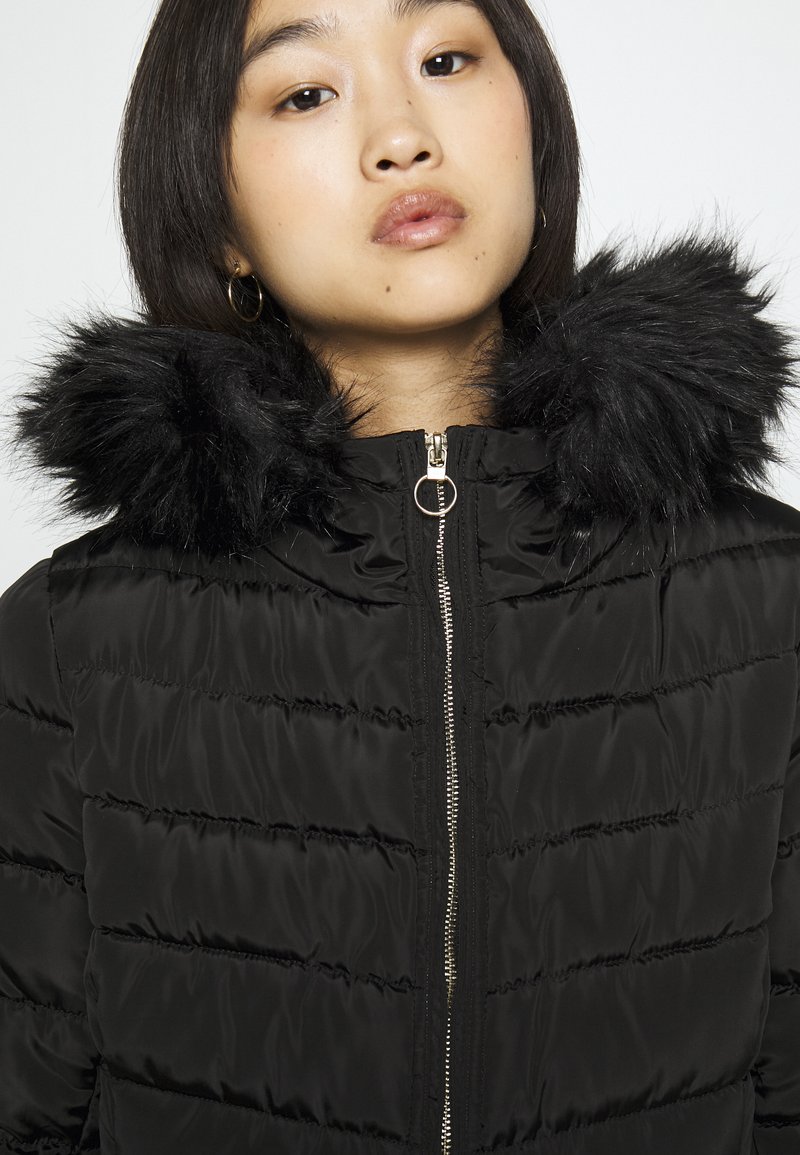 Giacca a vento nera con chiusura con zip e cappuccio in pelliccia sintetica. Presenta cuciture orizzontali e una superficie in tessuto liscia e lucida.