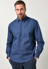 Hombre con barba que lleva una camisa de manga larga de patrón azul marino con botones y pantalones oscuros, con las manos en los bolsillos frente a un fondo liso.