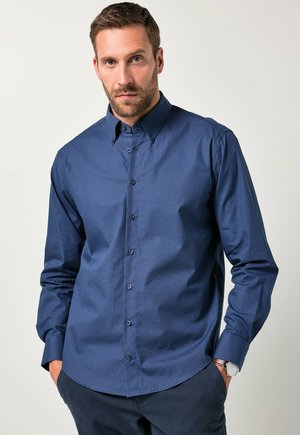Man met een baard, gekleed in een donkerblauwe gebloemde blouse met lange mouwen en donkere broek, met de handen in de zakken tegen een effen achtergrond.