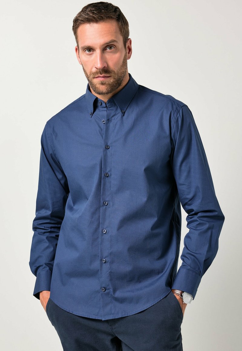 Hombre con barba que lleva una camisa de manga larga de patrón azul marino con botones y pantalones oscuros, con las manos en los bolsillos frente a un fondo liso.