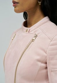 Svetlo roza umetni semisužni jopič z ovratnikom, zlatimi zadrgami in rebrastimi detajli na ramenih. Prisotno je podrobno šivanje.