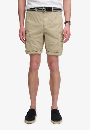 Uomo che indossa pantaloncini cargo beige con risvolti arrotolati, cintura nera, camicia bianca, orologio da polso nero e scarpe slip-on nere su sfondo bianco.