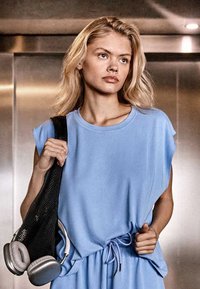 Jeune femme en haut bleu sans manches décontracté et pantalon porte un sac noir avec des écouteurs gris, debout devant un fond métallique.