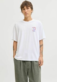 Jauns vīrietis ar auskariem, tetovējumiem uz abām apakšdelma pusēm, ģērbies baltā lielā T-kreklā ar maziem uzrakstiem un pelēkās brīvās biksēs, stāv uz vienkārša fona.