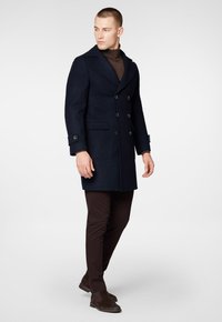 Granatowy wełniany płaszcz dwurzędowy z dużymi klapami, kieszeniami bocznymi i czarnymi guzikami, w zestawieniu z brązowym golfem i ciemnymi spodniami.