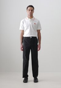 Chemise polo blanche à manches courtes avec des accents rouges, associée à un pantalon noir et des chaussures noires. Logo Lacoste affiché sur la chemise.