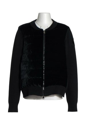 Sort lynlåsjakke med quiltet forpanel og strikkede ærmer vist på en hvid torso mannequin mod en ensfarvet baggrund.