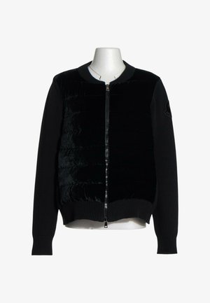 Sort lynlåsjakke med quiltet forpanel og strikkede ærmer vist på en hvid torso mannequin mod en ensfarvet baggrund.