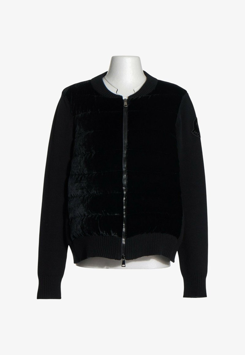 Veste noire zippée avec panneau avant matelassé et manches tricotées, présentée sur un mannequin torse blanc sur fond uni.