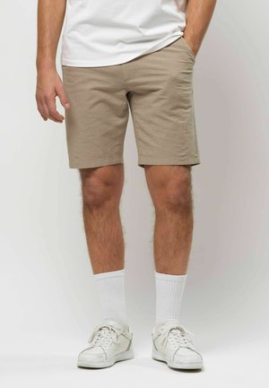 Muž oblečený v béžových rebrovaných šortkách, bielych členkových ponožkách, bielych teniskách a bielom tričku, stojaci s jednou rukou v vrecku.