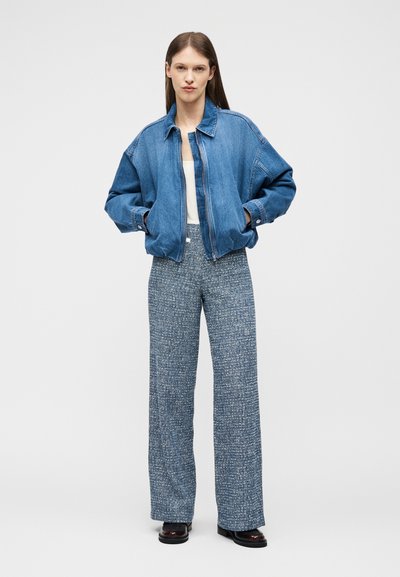 Femme debout, les mains dans les poches, portant une veste en denim bleue, un haut blanc, un pantalon gris à jambe large et texturé, ainsi que des chaussures foncées sur fond blanc.