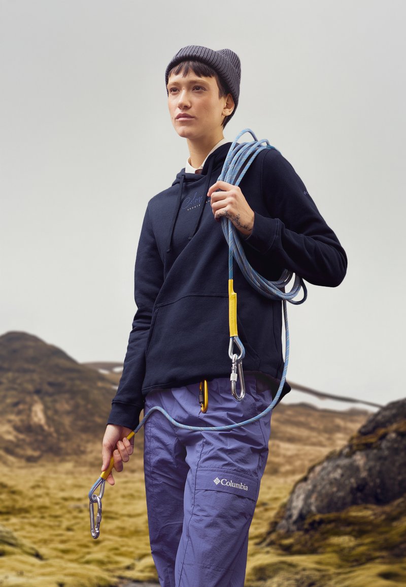 Felpa nera con logo, berretto grigio, pantaloni viola, che tiene una corda da arrampicata blu con dettagli gialli e moschettoni, ambientato su un paesaggio roccioso.
