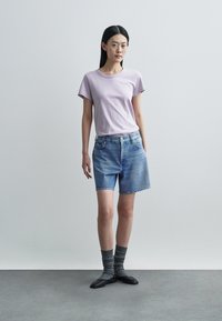 T-shirt en coton violet, short en denim bleu clair à coupe décontractée, chaussettes grises texturées et chaussures plates noires, le tout sur un fond neutre.