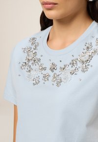 T-shirt bleu clair à manches courtes avec des embellissements floraux ornés de perles transparentes et argentées autour de l'encolure. Matière en coton douce.