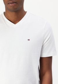 Mann trägt ein schlichtes weißes V-Neck T-Shirt mit kleinem Tommy Hilfiger-Logo in Rot, Weiß und Blau auf der linken Brust.