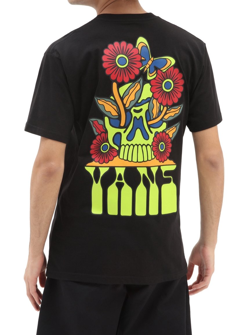 Personne portant un t-shirt noir avec un crâne coloré, des fleurs rouges, un papillon bleu et jaune, et un grand texte vert "VANS" dans le dos.