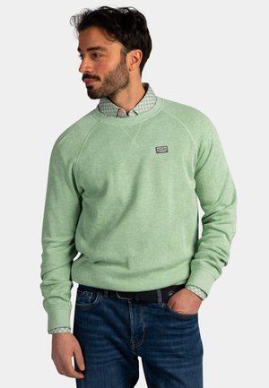 Man die een lichtgroene trui draagt over een overhemd met patroon en blauwe spijkerbroek, staat met één hand in zijn zak en kijkt naar links.