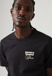 Juodas medvilnės marškinėlis su baltu Levi's logotipu ir "California" užrašu kairėje krūtinės pusėje; apvalus kaklas ir trumpomis rankovėmis.