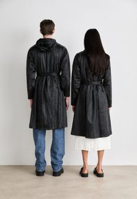 Two figures in black, long, hooded coats with belted waists; one in denim and the other in a white skirt, both wearing dark footwear.