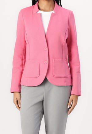 Kobieta w dopasowanej różowej marynarce z dwoma guzikami i naszywanymi kieszeniami, na białym topie i jasnoszarych spodniach.