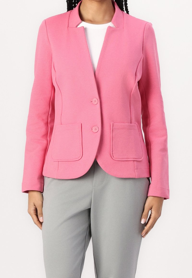 Femme portant un blazer rose ajusté avec deux boutons et des poches plaquées sur un top blanc et un pantalon gris clair.