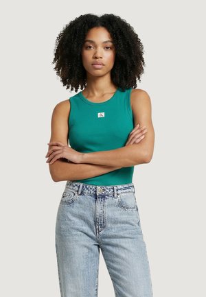 Mujer con cabello rizado, vestida con una blusa sin mangas color verde azulado y jeans de tiro alto azul claro, de pie con los brazos cruzados sobre un fondo liso.